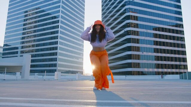 Fashionable funky african american girl dancer in orange sportswear performing outdoor street dancing motion at urban background. Joyful female artist demonstrate choreography in city golden sunset 4K