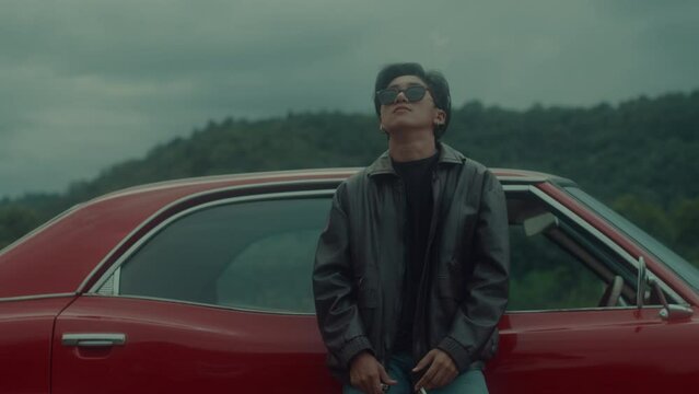 A brutal tomboy girl in a brown leather jacket leans on her vintage red car and smokes