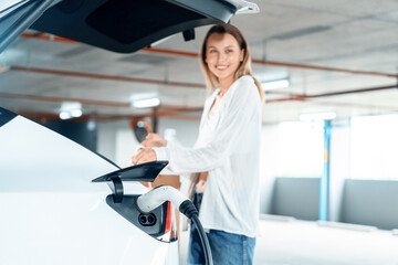 Young woman travel with EV electric car to shopping center parking lot charging in downtown city...