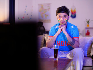 An Indian man in a blue cricket jersey watching a live cricket match on TV or television - cricket...
