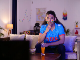 Obraz premium Excited young Indian man with long hair wearing cricket jersey watching cricket match on tv excitement. An excited Indian cricket fan - watching sports excited live tv ott platform cricket se...