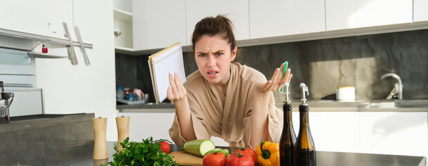 Portrait of woman cant cook, looking confused while making meal, holding recipe book, checking grocery list and staring frustrated at camera, standing near vegetables in the kitchen - obrazy, fototapety, plakaty