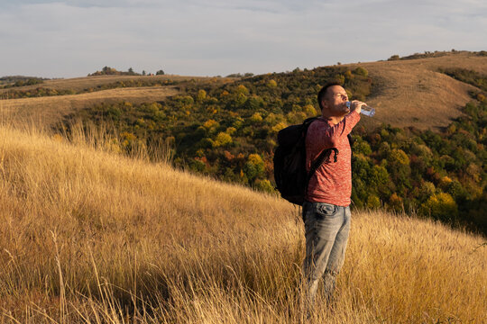 Young man drinking water after hiking through autumn fields and hills against blue sky during warm day. Weekend, leisure, sport