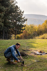 Naklejka na ściany i meble Male tourist chopping firewood for bonfire with axe in meadow after hiking during warm sunny autumn day. Survival, travel, hiking, camping in nature