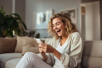 Deurstickers Happy excited young woman relaxing on couch using mobile phone winning in online app game. Young lucky girl feeling winner looking at cellphone, receiving great news or discount offer. © radekcho