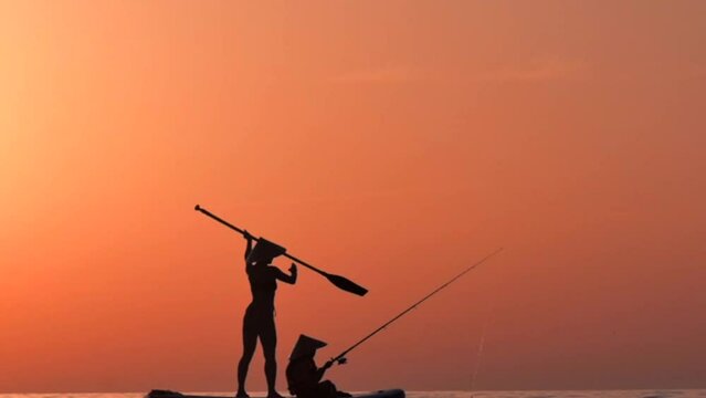 girl and a child on sup boards swims to the sunset on the sea with a fishing rod