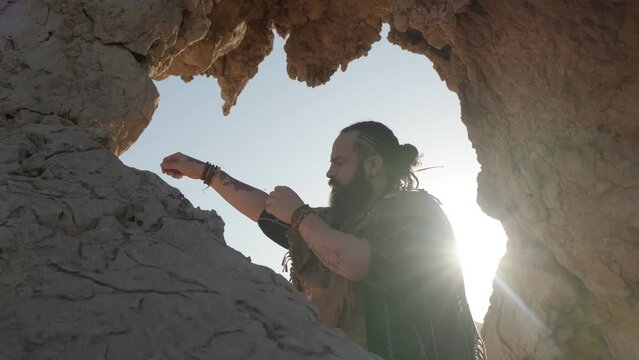 A young bearded shaman prays while standing in a grotto of a rock among the sand dunes of the desert