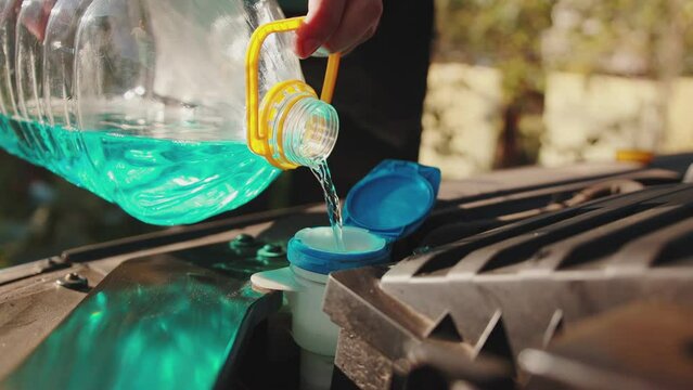 A man opens the cap of the windshield washer fluid reservoir and pours in blue anti-freeze liquid for cleaning glass. Slow motion, close-up