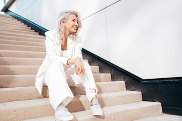 Young beautiful blond woman wearing nice trendy white suit jacket. Smiling model posing in the street at sunny day. Fashionable female outdoors. Cheerful and happy. Sits at the stairs