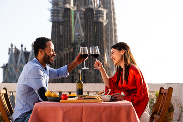 Multiracial beautiful happy couple of lovers dating on rooftop balcony at Sagrada Familia, Barcelona - Multiethnic people having romantic aperitif dinner on a terrace with city view 