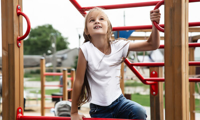 Fototapeta premium A playful little baby girl climbed an iron ladder on an outdoor playground. A cheerful preteen girl climbs the stairs on the playground. Games for the development of children outdoors.