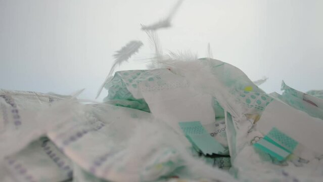 Feathers fall on a stack of baby diapers on a white background. The concept of softness and comfort, thorn-allergenic diapers made of cotton. Copy space for text. Slow motion