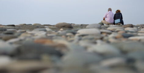 back, rear view of Senior couple sitting on stone pebbles beach looking at distance. pensioner pair sitting on Black Sea coast. Senior mature couple from behind on summer or autumn beach.