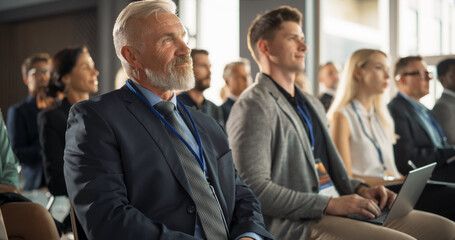 Senior Caucasian Male Entrepreneur Sitting In Diverse Crowd On Business Forum. Successful Investor Listening To Presenter Pitching a Startup Idea On Stage. Businessman Attending Technology Conference.