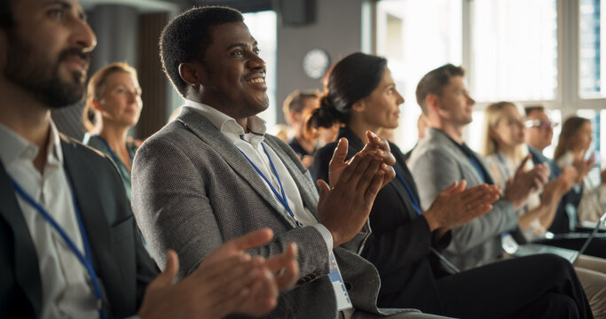 African Man Sitting in Crowded Auditorium at a Tech Conference. Black Businessman Listening To Interesting Keynote. Specialist Watching Innovative Technology Presentation And Clapping With Audience.