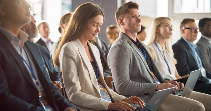 Young Woman Sitting in a Crowded Audience at a Business Conference. Female Attendee Writing Important Information on a Laptop. Keynote Speech in an Auditorium with Successful Business Executives.