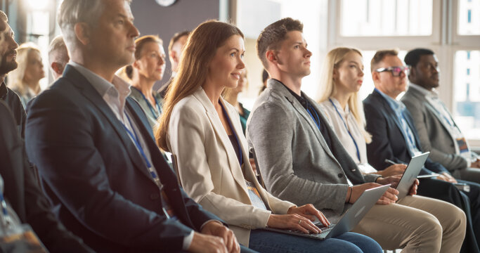 Young Woman Sitting in a Crowded Audience at a Business Conference. Female Attendee Writing Important Information on a Laptop. Keynote Speech in an Auditorium with Successful Businesspeople.