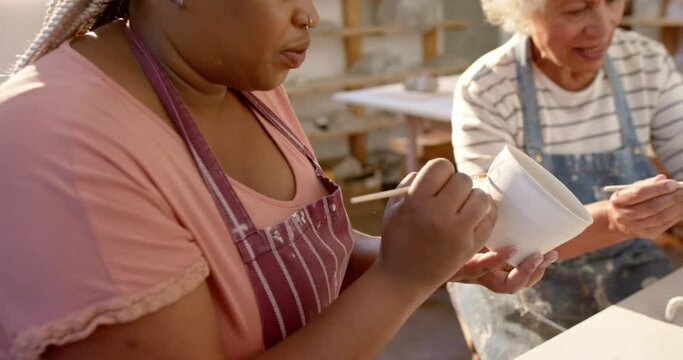 Two diverse female potters glazing clay jug and smiling in pottery studio, slow motion