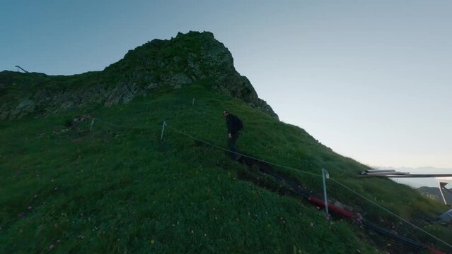 Male backpacker climbing on stairs rope summer morning mountain summit sunny ski resort aerial view. Travel man hiker going upstairs on natural cliff rock covered green grass effort journey adventure