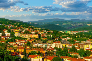panoramic landscape view of green summer city with yellow houses, green forest and mountains above...