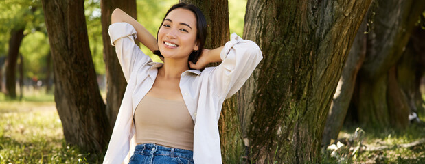 Portrait of beautiful asian woman resting near tree, relaxing in park, smiling and looking happy