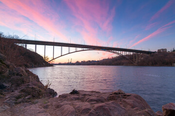 Steel arched bridge on Old Dnieper with bright clouds in evening sky reflecting in water and ancient granite rocks, Khortytsia island, Zaporizhzhia, Ukraine