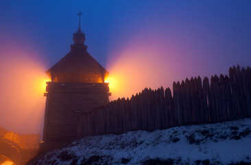 Wooden tower of cossacks fortress "Zaporizhian Sich" in evening fog with rays of orange illumination lights, Khortytsia island, Zaporizhzhia, Ukraine