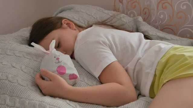 A little girl sleeps on a bed during the daytime, putting her hand on a gift bag with a rabbit filled with sweets for Easter or Christmas. Happy childhood.