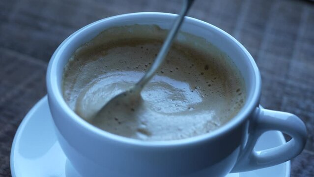 Stirring thick foam of hot coffee cappuccino with spoon in white cup on wooden table, close up. Milk foam cappuccino, latte