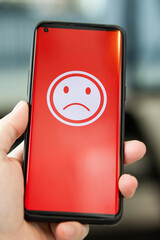 A woman holding a smartphone with a bad feeling emoticon on screen at a restaurant. concept of...