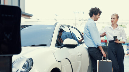 Progressive businessman and businesswoman leaning on electric car connected to charging station...