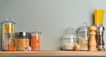 Various uncooked groceries and pasta in glass jars arranged on wooden shelf on olive green wall.