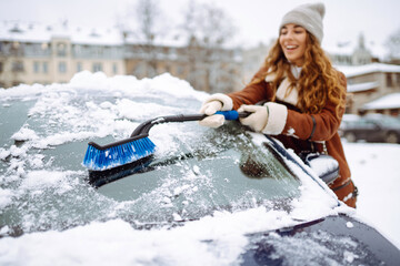 Time to clear the snow! A young woman cleans snow from a car with a brush. Winter car concept.