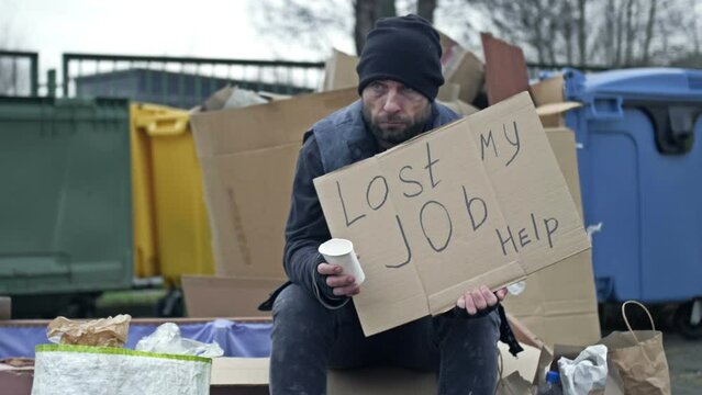 Cold and dirty young man sits by a pile of rubbish and eats and drinks something. Next to it is a handwritten LOST MY JOB HELP poster.