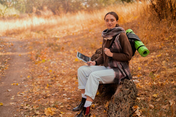 Fototapeta premium girl with a backpack and a map in the forest, sitting on a log, looking at a map in the autumn forest, autumn holidays and hiking, lifestyle concept