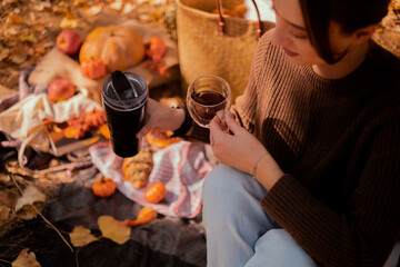 Woman pouring tea from thermos into cup relaxing in autumn forest sitting on trunk. Close up of hot drink and basket with yellow leaves