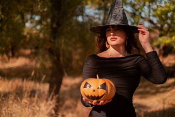 Beautiful woman in witch costume with Jack-o'-lantern in forest, Halloween party, smiling evil...