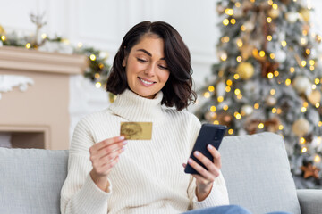 A young beautiful woman is sitting on the sofa at home, holding a credit card and phone and doing online shopping on the eve of the New Year and Christmas holidays