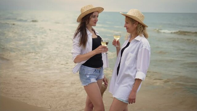 Two young beautiful hipster woman in trendy summer shirts and hats drinking wine on sea picnic. Having picnic on summer evening