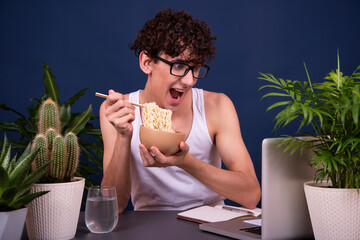 A funny young man in a white T-shirt eats noodles and teaches online lectures. A guy on a blue...
