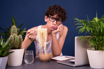 A funny young man in a white T-shirt eats noodles and teaches online lectures. A guy on a blue...