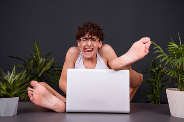 A funny young man sits at a laptop with his feet up on the table. Watching videos for adults. Dating on the Internet. - obrazy, fototapety, plakaty