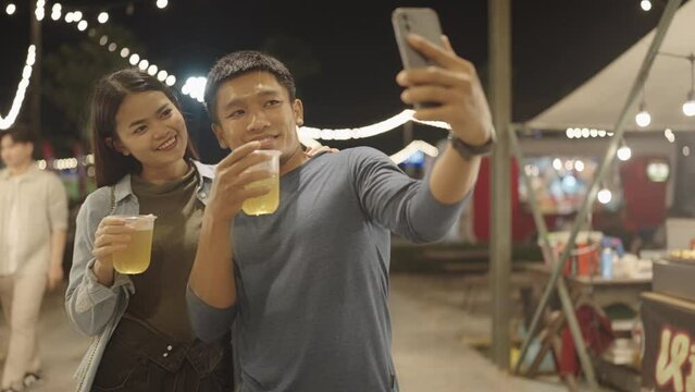 Happy young Asian couple drinking beer alcohol and taking selfie with smartphone in night market