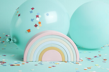 a plastic rainbow toy in front of balloons of the same color as the background and confetti on the ground.