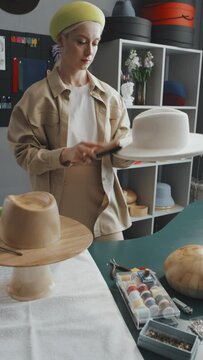 Vertical shot of young hat-maker standing in her workshop while brushing out new white felt hat