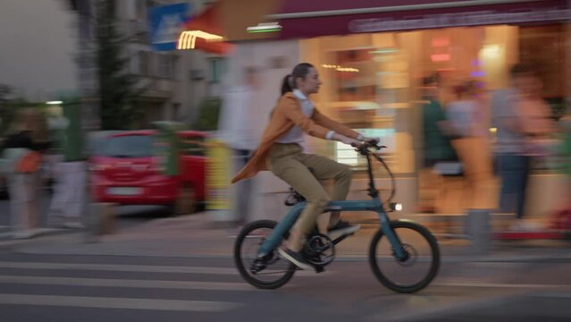 Stylish young woman riding an electric bicycle in city centre in modern street during fall season. Concept of urban cycling and eco transportation by bike