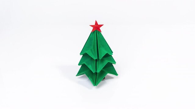 An origami Christmas tree is folded from green paper on a white background. Red star decorates the top of the spruce tree. Original Greting Card. Copy space. Stop motion animation.