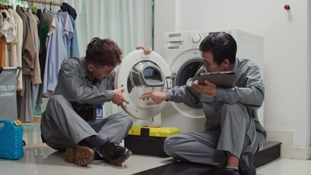 Medium long shot of two Asian plumbers looking at instruction on digital tablet while installing washing machine in clients house