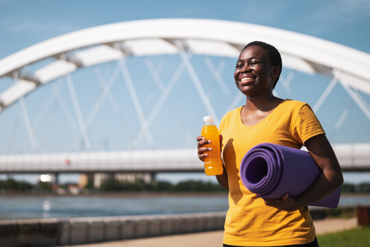 Sporty woman drinking energy drink and holding exercise mat.	