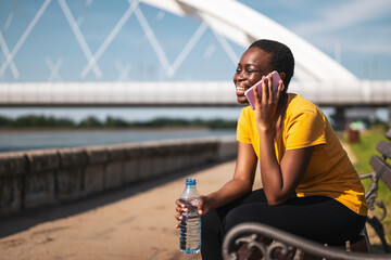 Woman with bottle of water using mobile phone while resting after exercise on bench.	